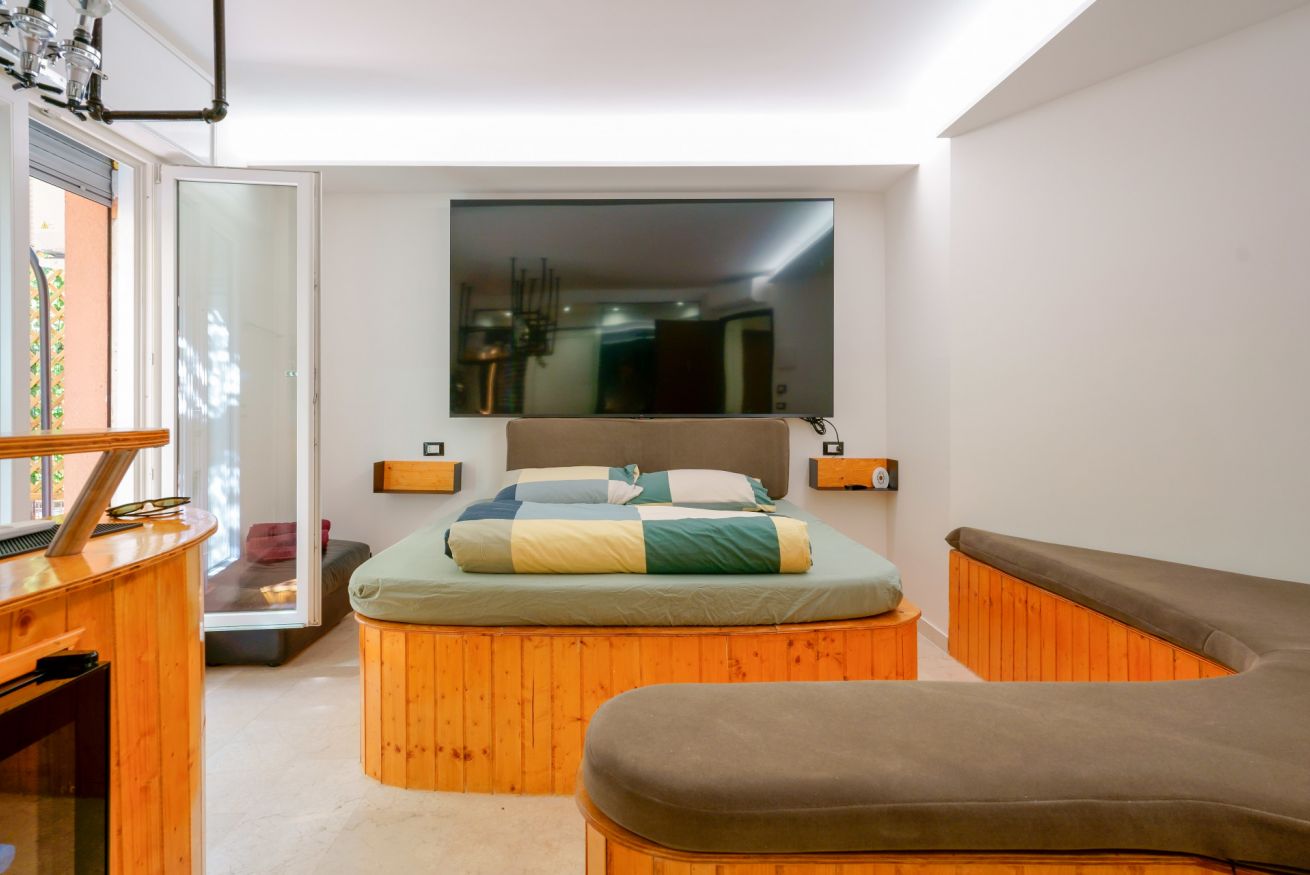 Camera matrimoniale con letto in legno naturale, arredo moderno e grande schermo sulla parete, illuminata da luce naturale e dettagli in arancio.