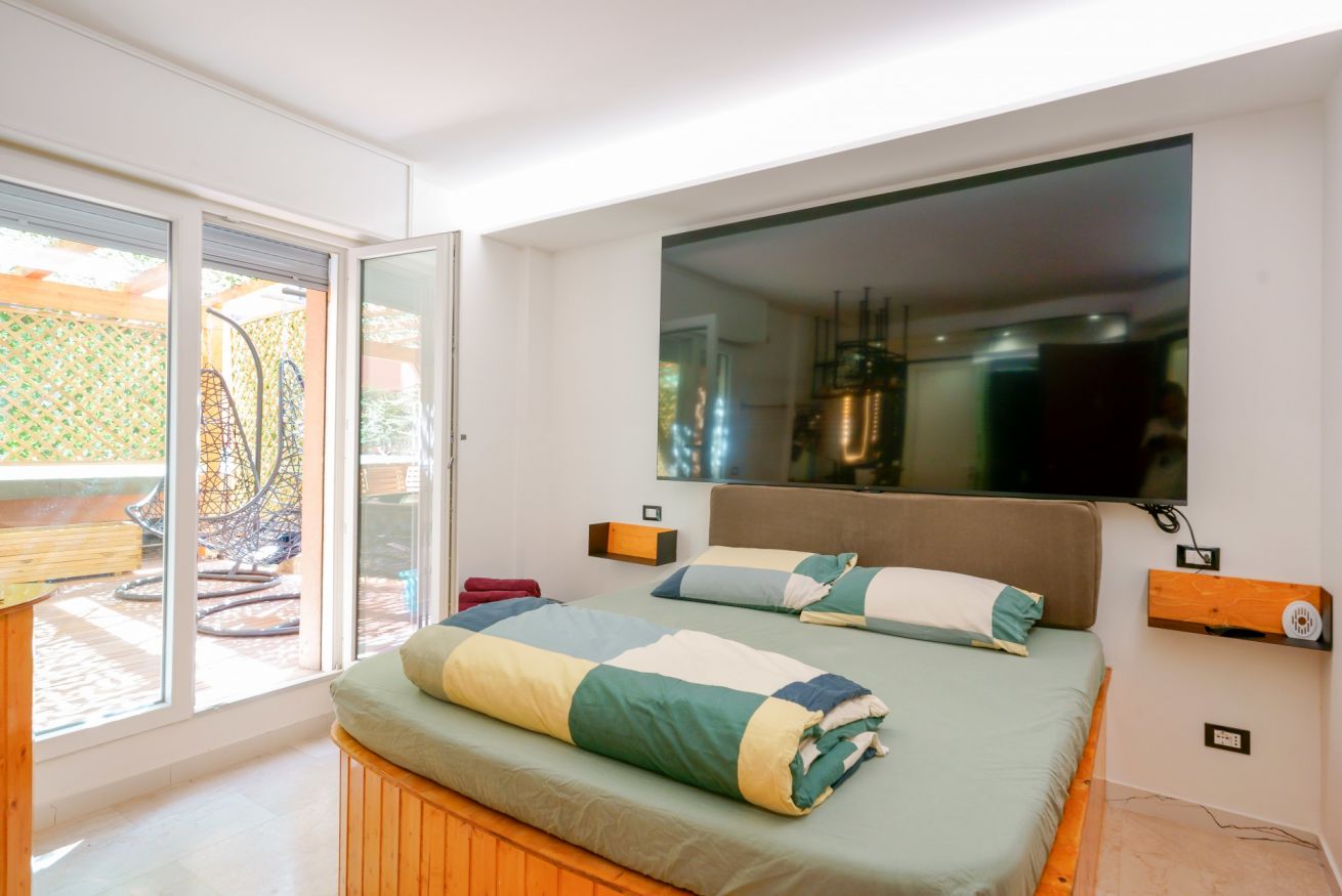Camera matrimoniale luminosa con terrazza privata, arredi moderni in tonalità naturali e accenti color arancio, grande schermo TV e accesso diretto all'esterno.