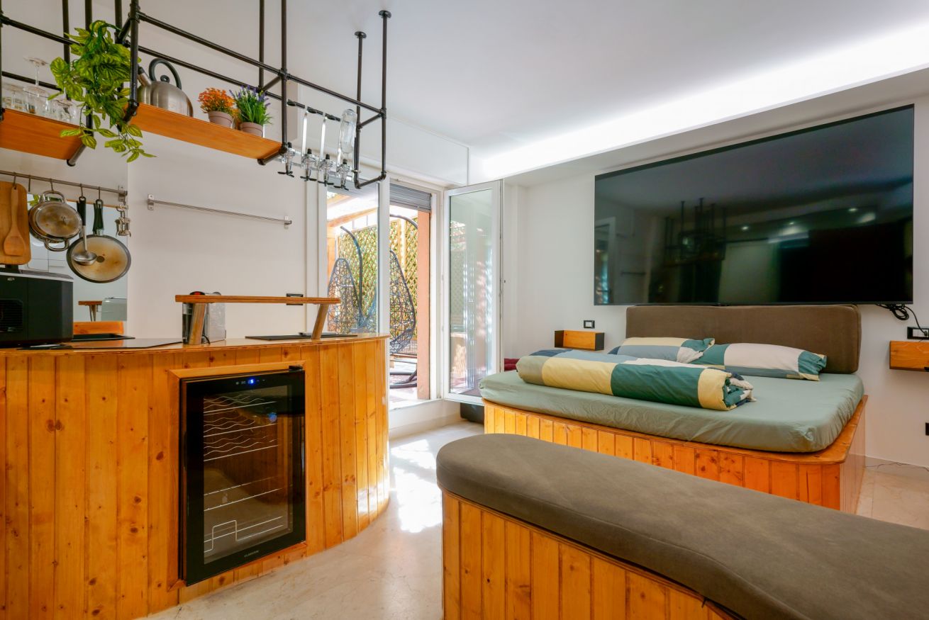 Elegante camera con letto matrimoniale su base in legno, zona living con kitchenette e accesso alla terrazza; design contemporaneo che unisce comfort e luminosità.