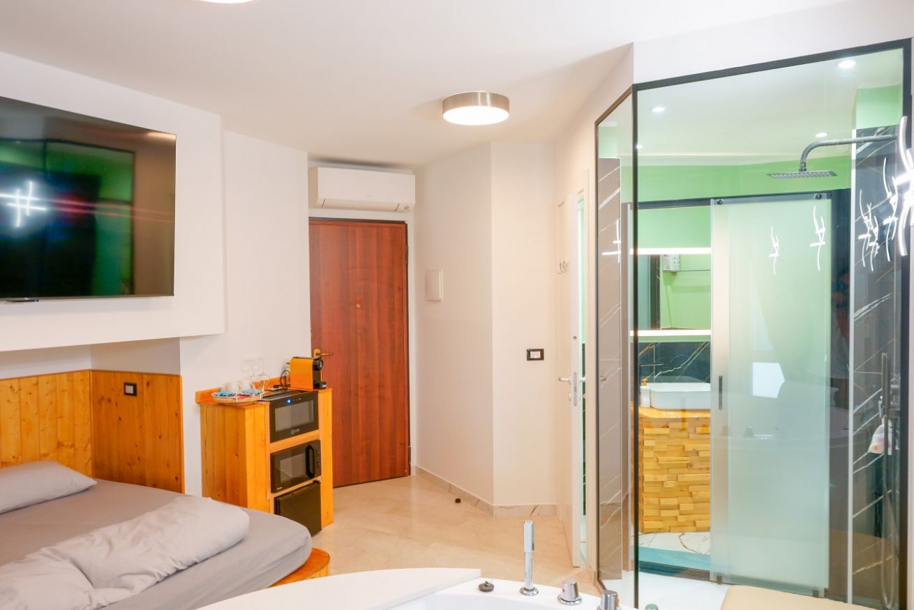 Camera moderna con letto matrimoniale, bagno con vasca idromassaggio in vetro, arredamento in legno caldo e TV a parete per il massimo comfort.