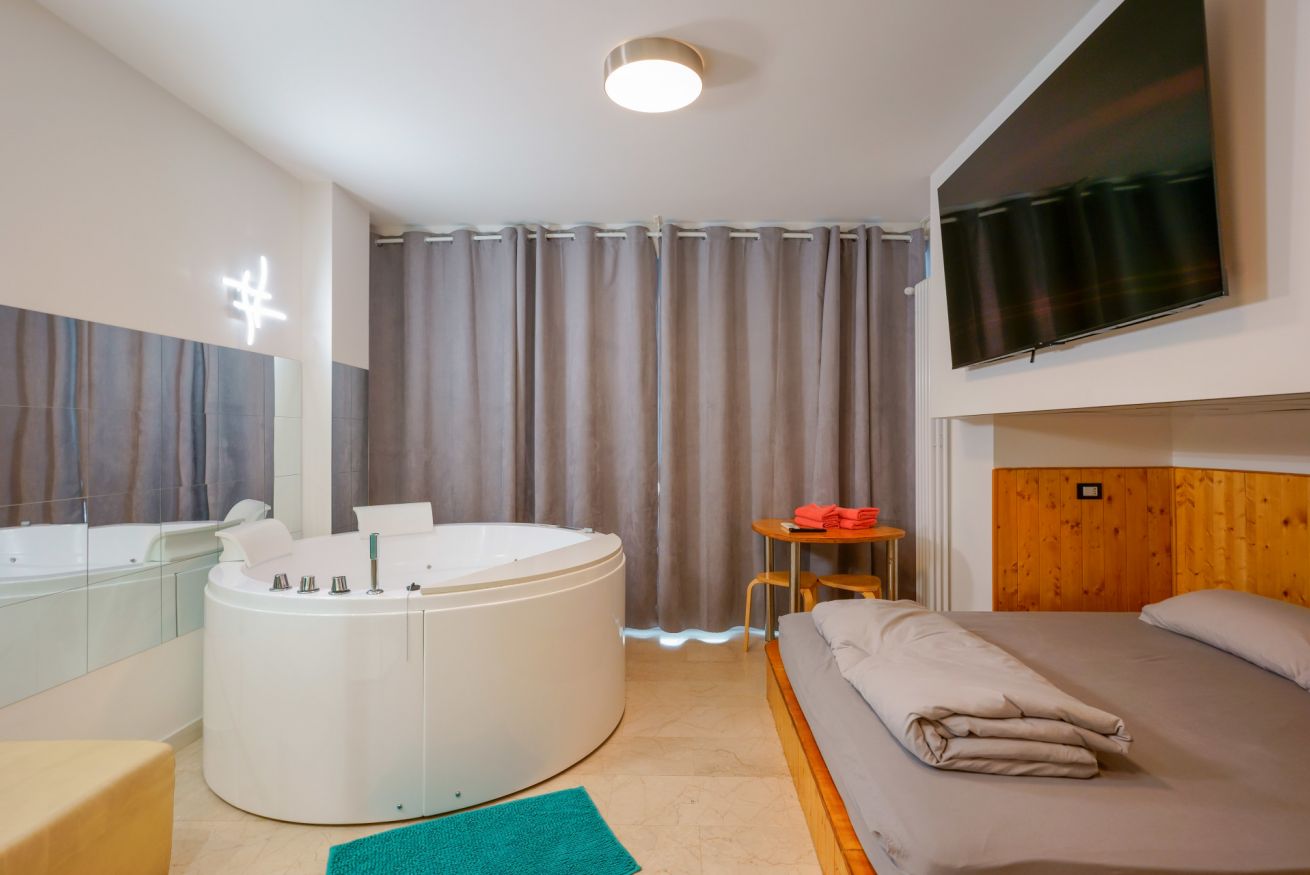 Camera accogliente con vasca idromassaggio circolare, letto matrimoniale in legno, TV moderna e tende eleganti per un soggiorno rilassante.