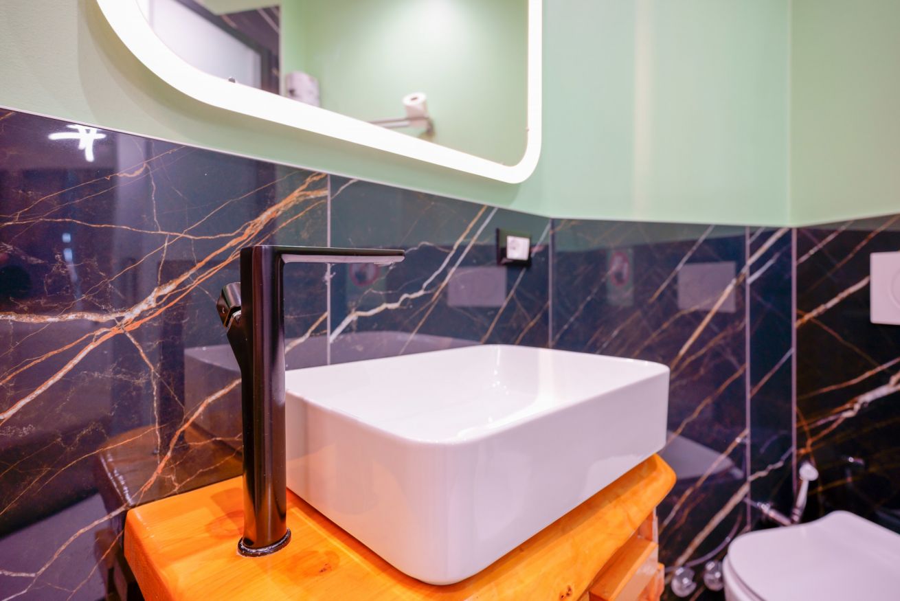Elegante bagno con lavabo moderno su base arancio, rivestimenti in marmo nero e specchio illuminato: un'oasi di lusso e relax.