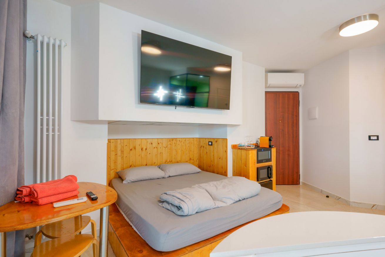 Accogliente camera matrimoniale con arredo in legno caldo, TV integrata sopra la testiera e ampi spazi luminosi per il vostro comfort.