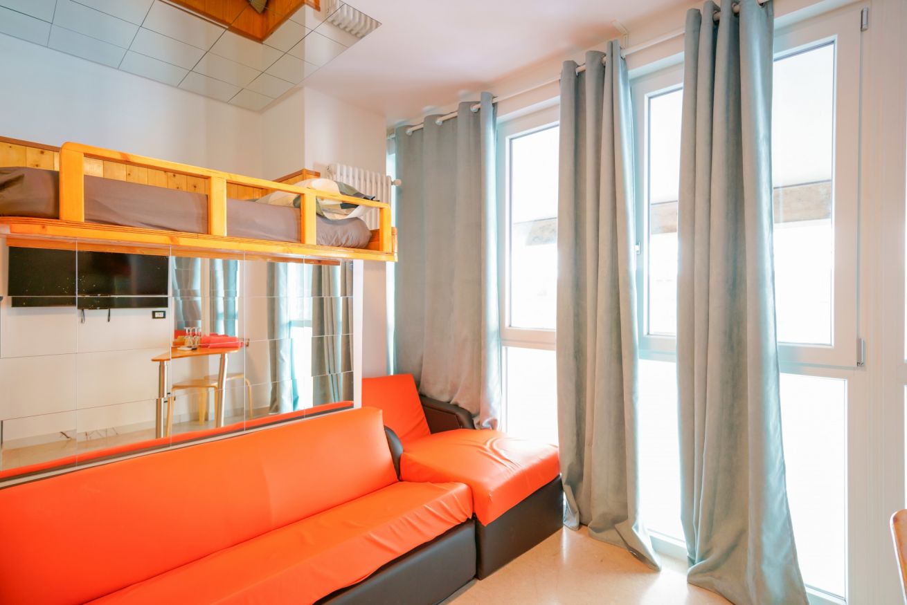Bright room featuring a vibrant red single bed, wooden mezzanine, and expansive windows overlooking the street, perfect for comfortable stays.