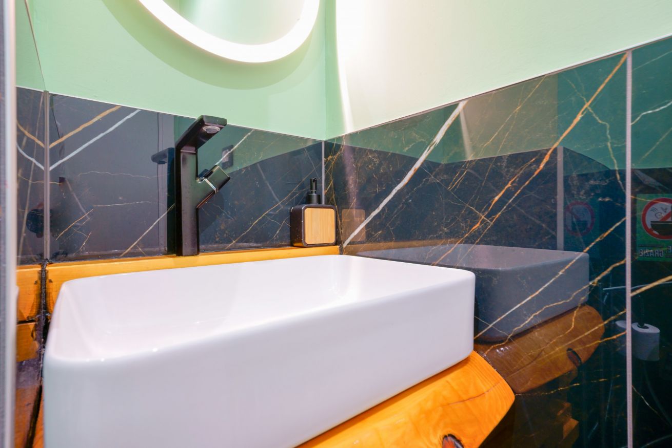 Bagno elegante con lavabo bianco, rivestimenti in marmo verde e dettagli in ottone, illuminazione moderna e design contemporaneo.