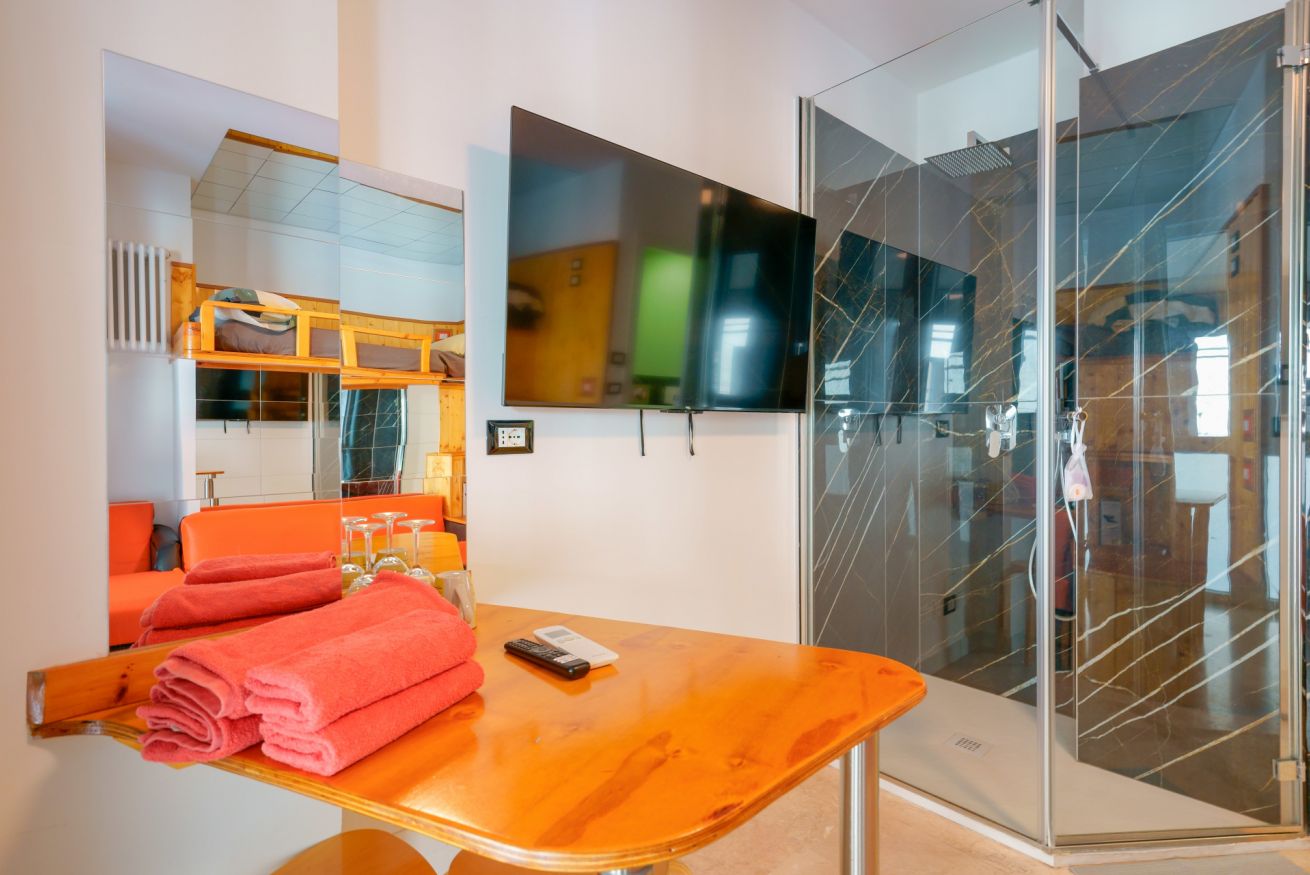 Camera singola con bagno privato in vetro, arredi moderni e tonalità calde, dotata di TV e linens di qualità.